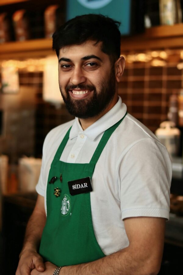 Friendly barista in green apron poses in a cozy coffee shop setting.